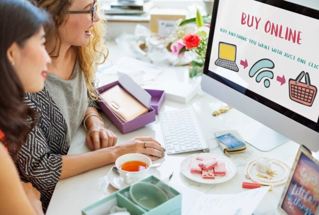 Two people are shopping online at a desk, featuring a computer screen with a Buy Online message and various items scattered around.