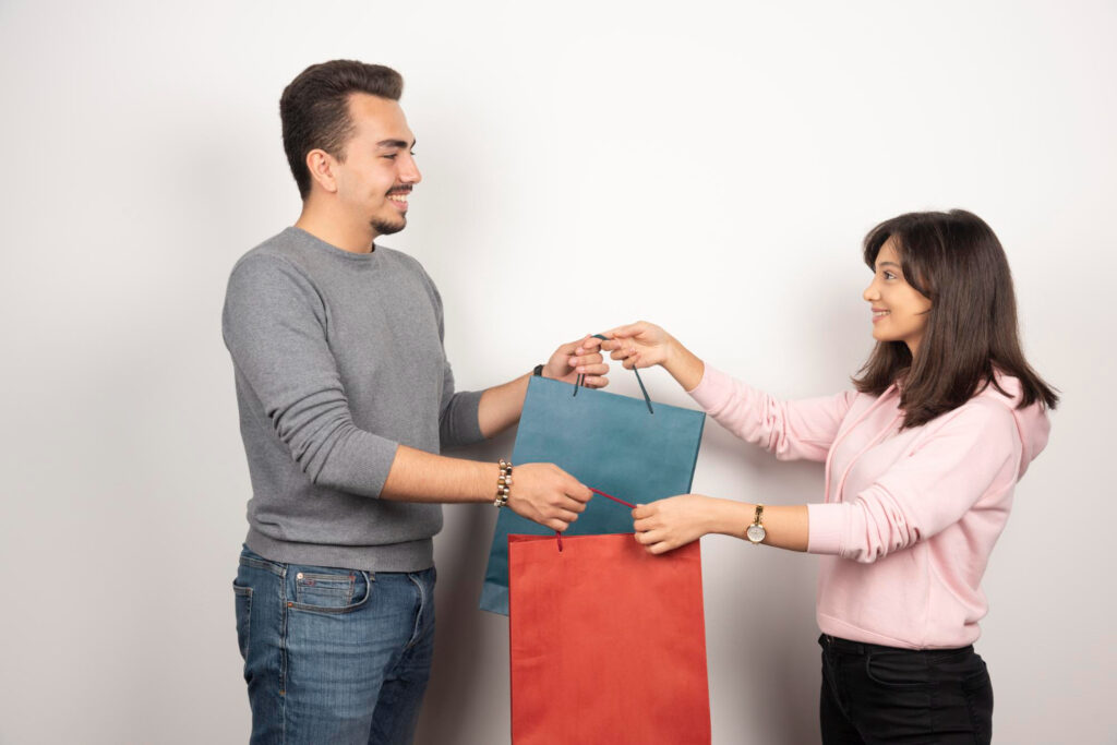 A man and woman exchange colorful shopping bags, smiling against a plain white background.