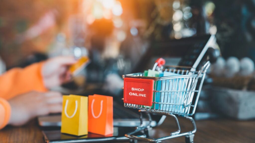 A person using a laptop for online shopping, with a miniature shopping cart labeled SHOP ONLINE and colorful paper bags nearby.