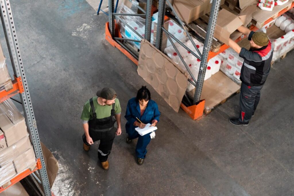 Two warehouse workers discuss while one writes on a clipboard, surrounded by shelves of boxed items and supplies.