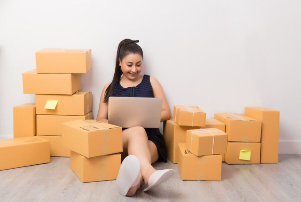 A person sits on the floor surrounded by stacked cardboard boxes, typing on a laptop, in a minimalist room.