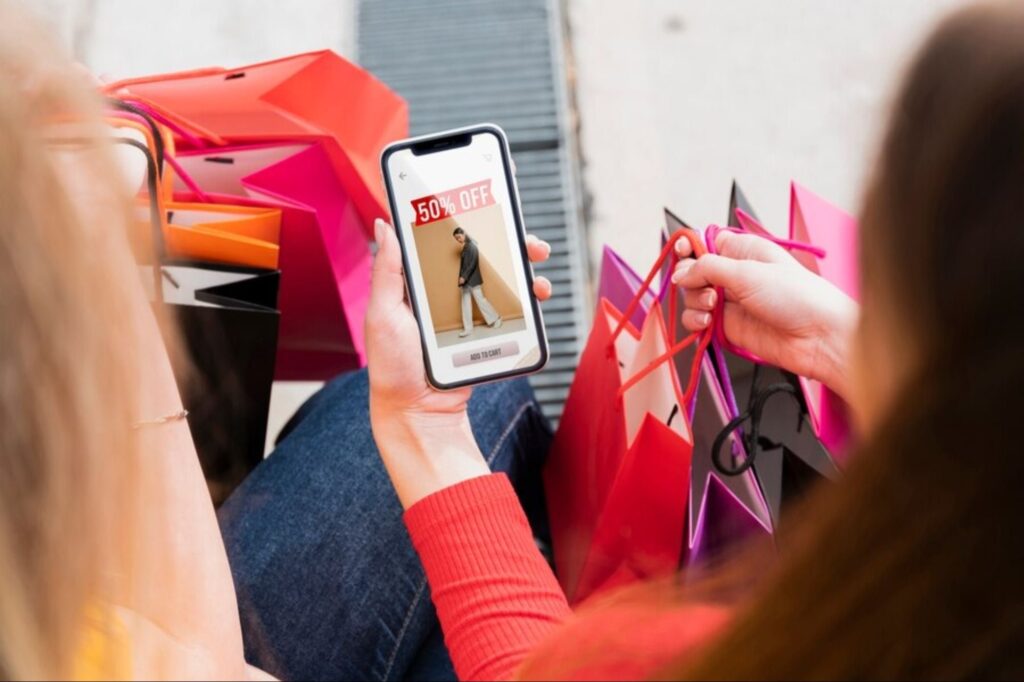 A person holding a smartphone displaying a 50% off sale, surrounded by colorful shopping bags.