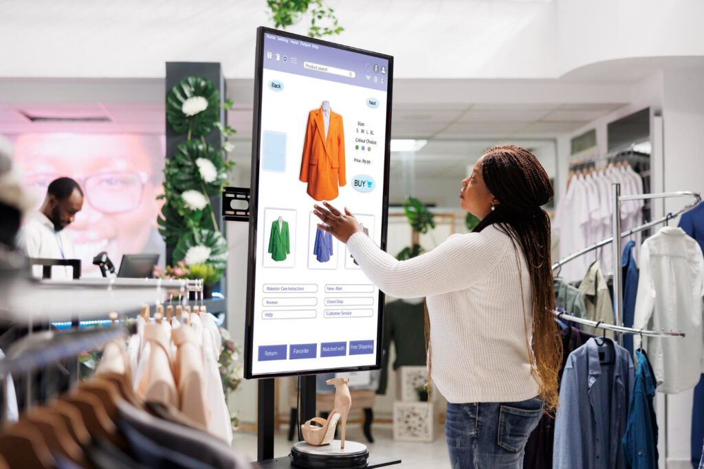 A shopper interacts with a digital screen displaying clothing options, including an orange blazer, green jacket, and blue skirt.
