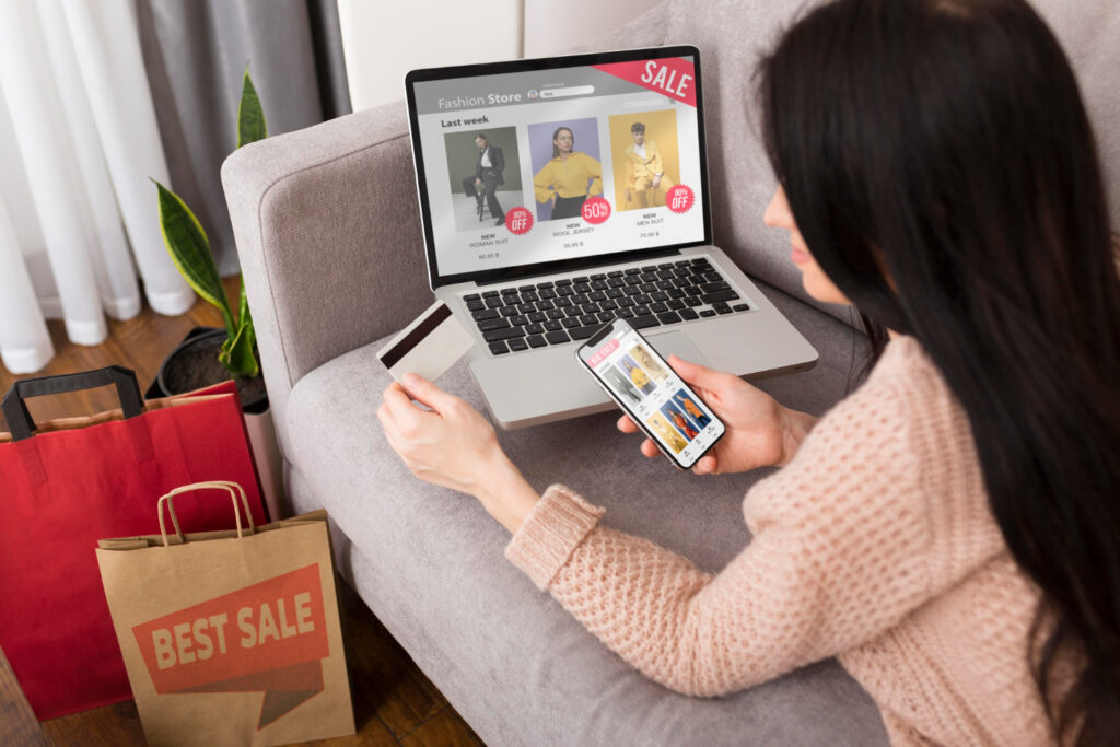 A person sits on a sofa shopping online for fashion items using a laptop and smartphone, surrounded by shopping bags.