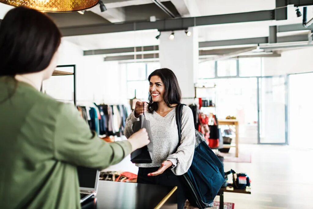 A customer is receiving a bag from a cashier at a retail store, surrounded by clothing racks and a modern, bright interior.