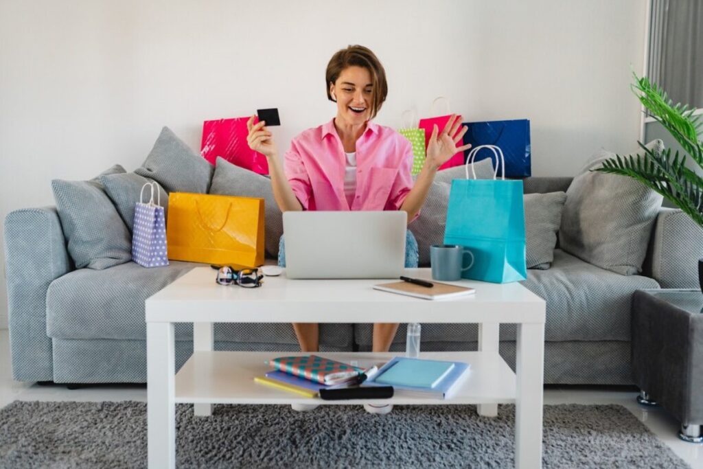 A person in a pink shirt sits at a table with a laptop, holding a shopping card, surrounded by colorful shopping bags.