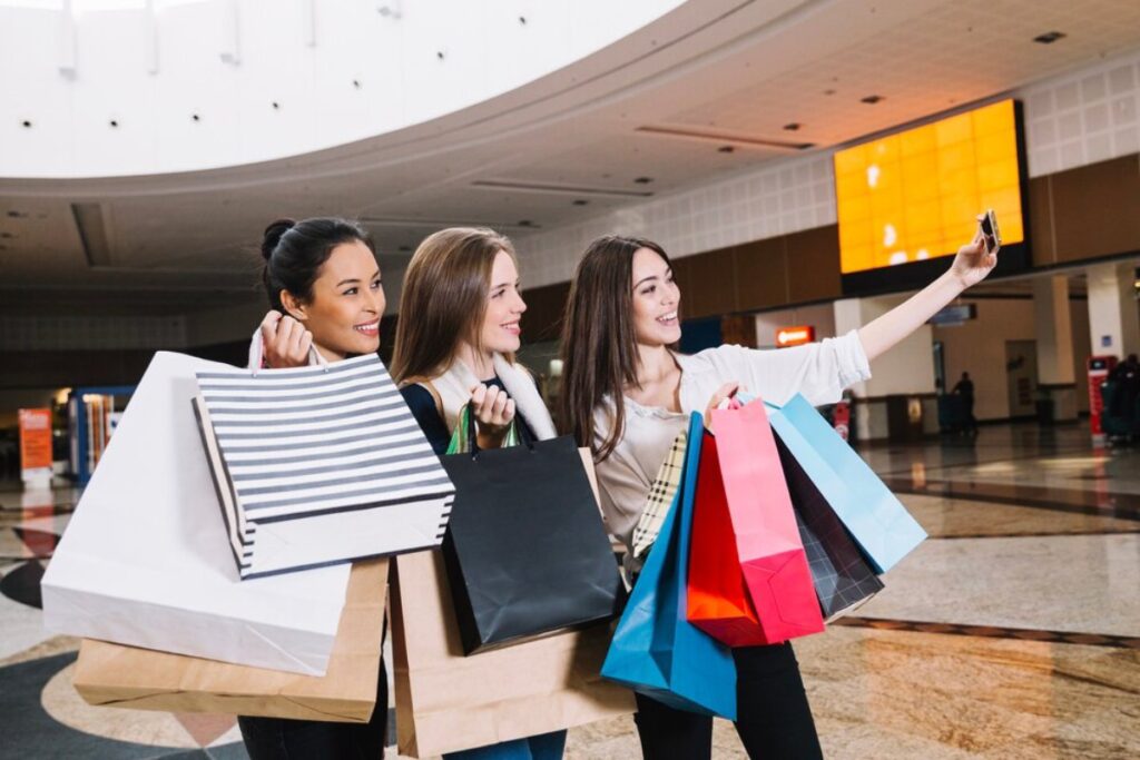 Three friends smiling and posing for a selfie while holding colorful shopping bags in a spacious indoor shopping mall.