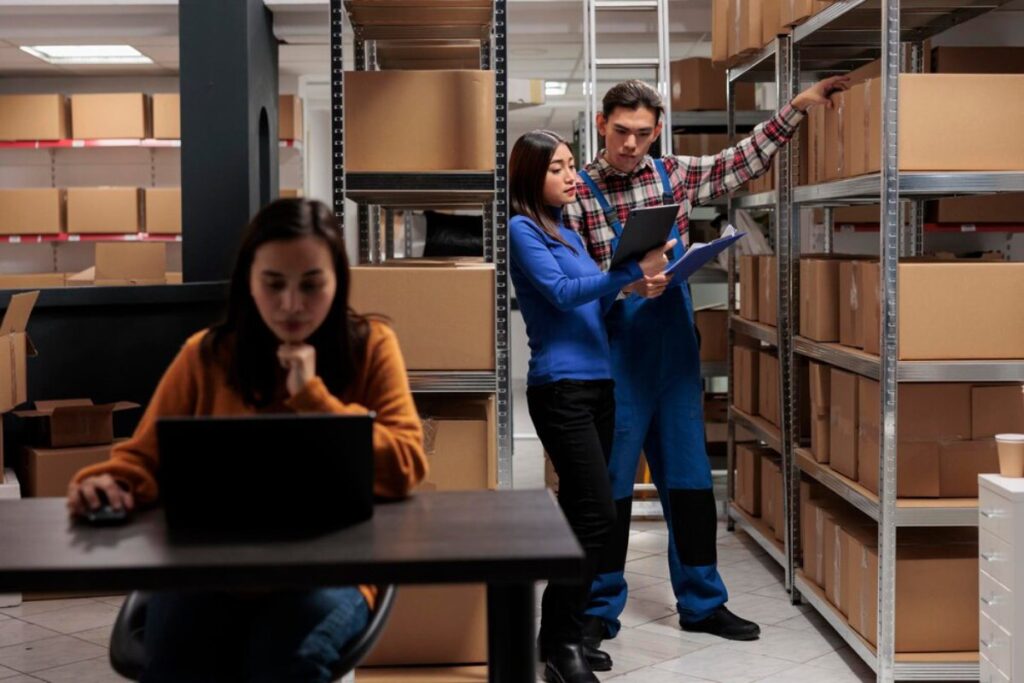 A woman sits at a computer while two colleagues discuss inventory in a storage area filled with cardboard boxes.