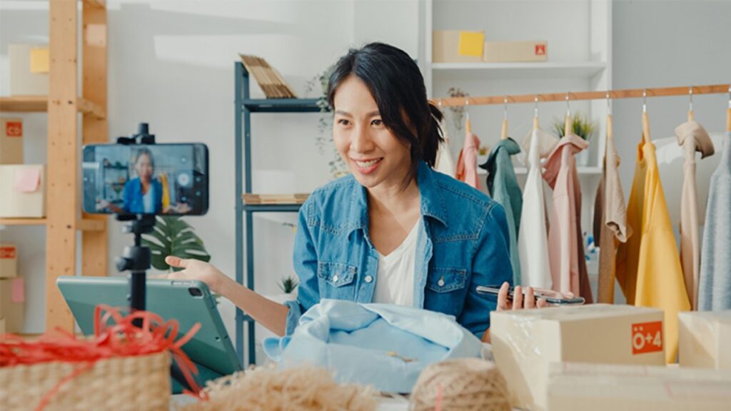 A person in a blue denim jacket gestures while recording a video, surrounded by clothing and packaging materials in a bright setting.