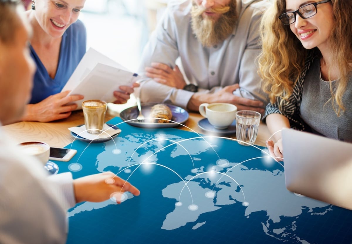 Four professionals discussing over a digital map on a table, with laptops, coffees, and smartphones.