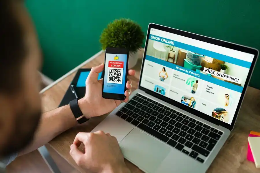 A person holds a smartphone displaying a QR code coupon while shopping online on a laptop with a Free Shipping banner.