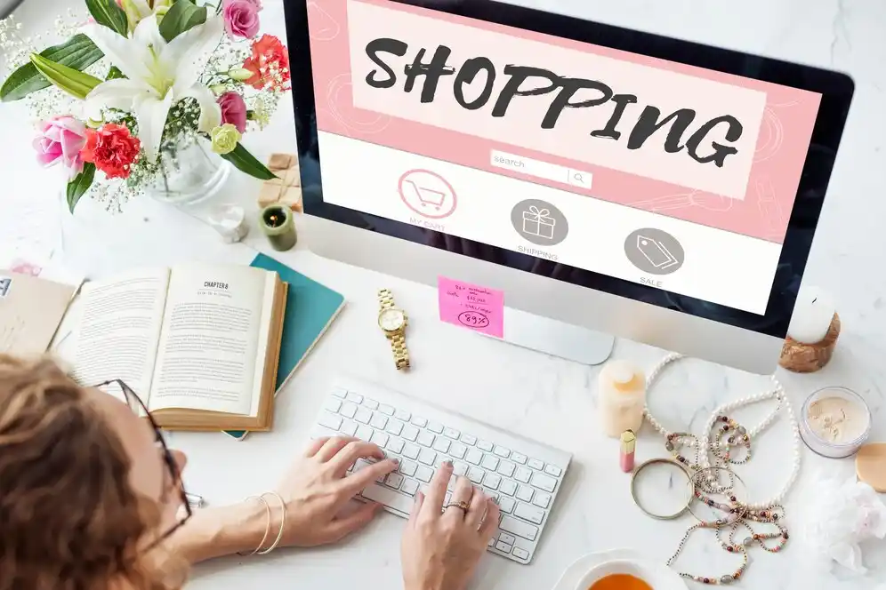 A hands-on keyboard in front of a computer displaying a vibrant shopping website, surrounded by flowers, an open book, and cosmetics.