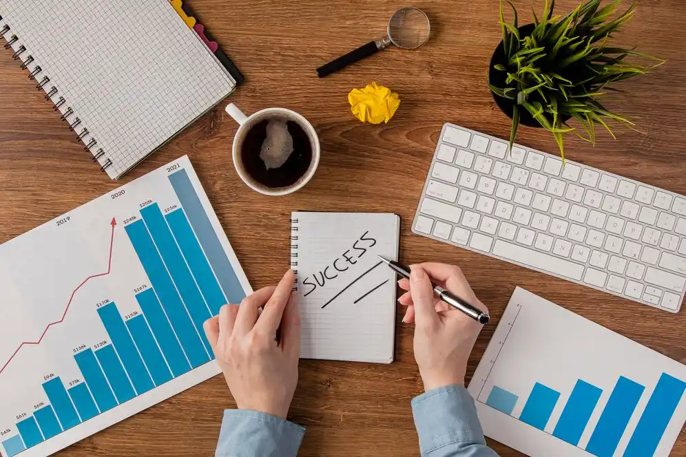 A person writes SUCCESS in a notebook surrounded by charts, a cup of coffee, a keyboard, and a small plant, symbolizing goal setting.