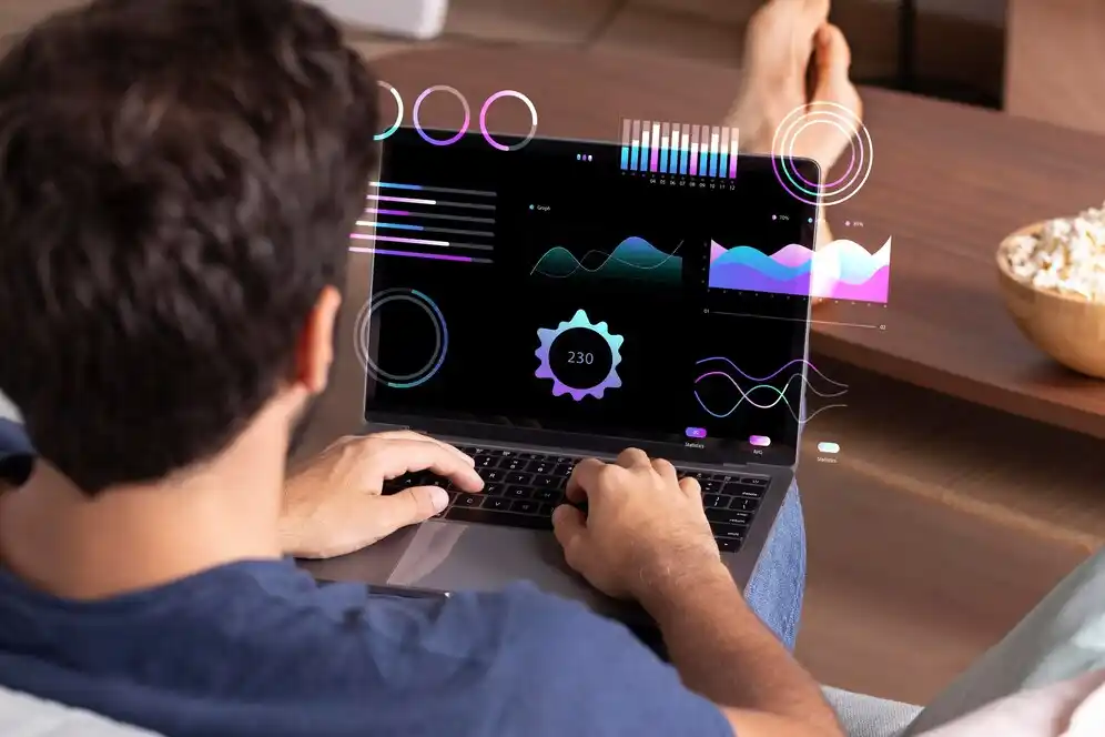 A person using a laptop with virtual data graphs and analytics displayed, while relaxing on a couch with a bowl of popcorn nearby.