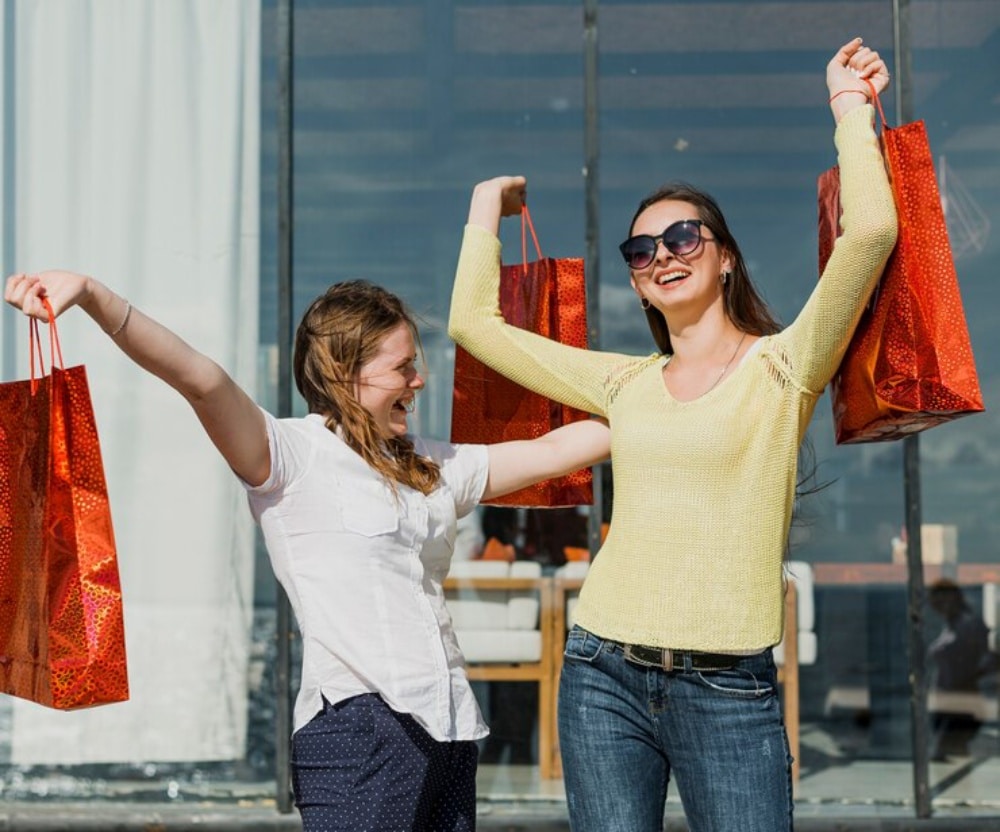 Two joyful women holding shopping bags aloft outside a store, enjoying a sunny day.