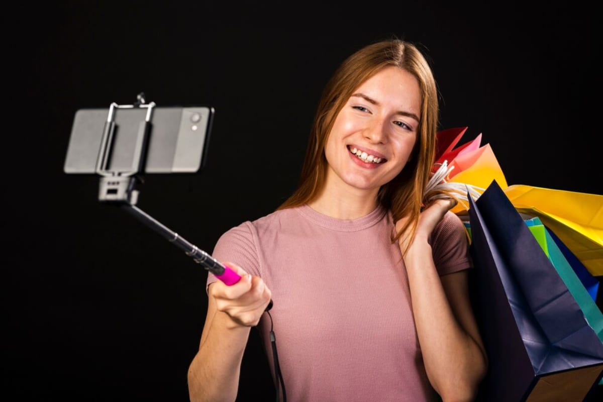 Young woman with shopping bags taking a selfie with a smartphone on a selfie stick, smiling, against a black background.