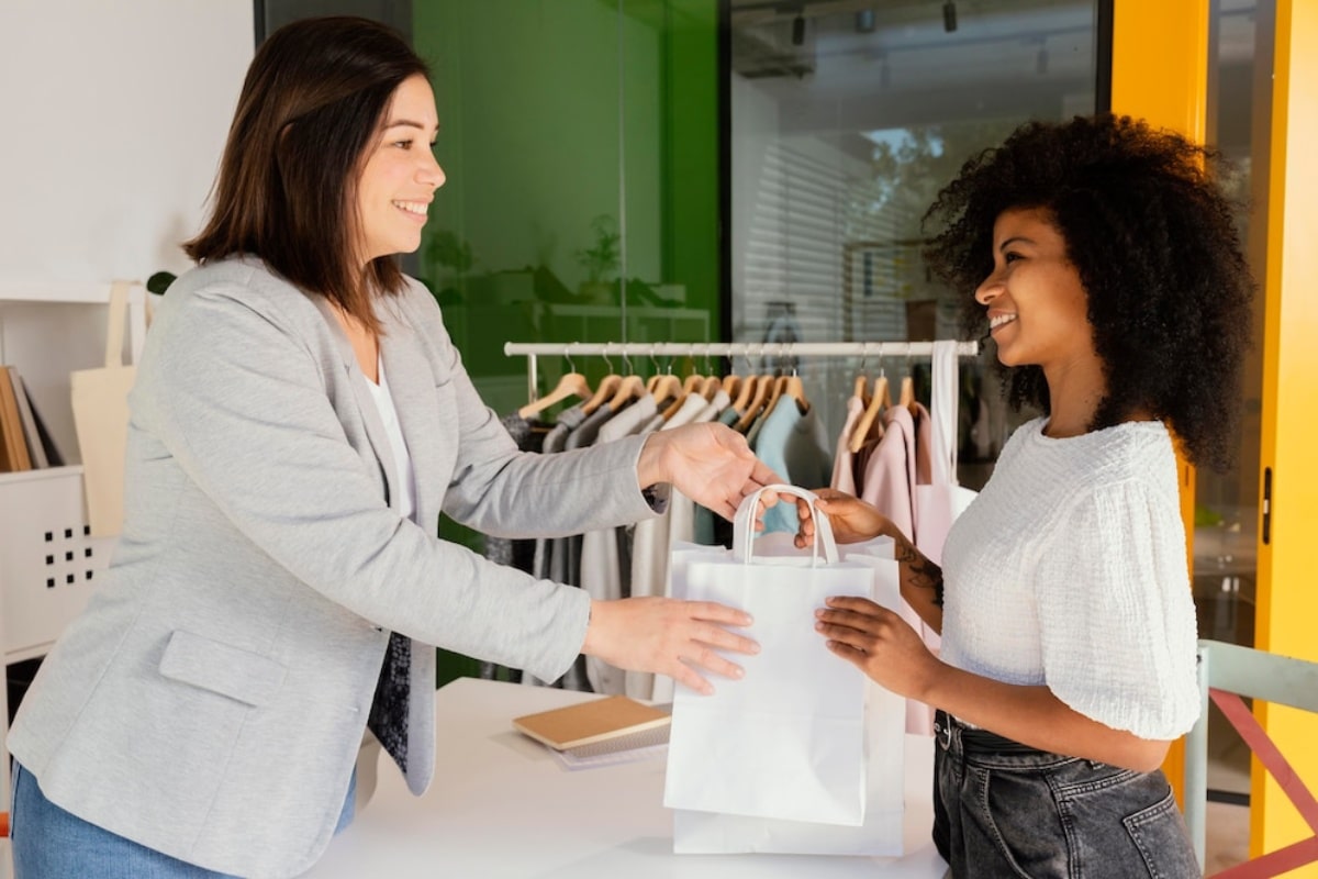 Two women smiling as one hands over a shopping bag to the other in a clothing store.