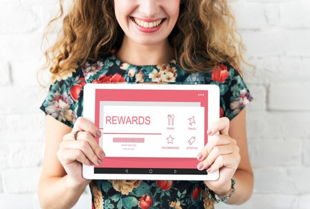 Person in floral dress smiling and holding a tablet displaying a rewards program interface against a white brick wall.