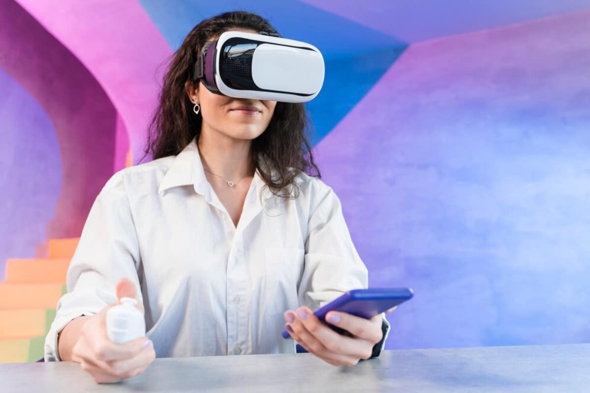 Woman wearing VR headset and holding a smartphone, sitting in a room with colourful geometric background.